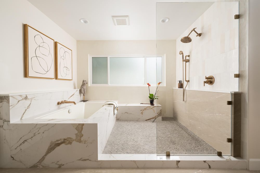 River stone pebble shower floor seamless glass door light oak vanity soft ambient ceiling spotlight.