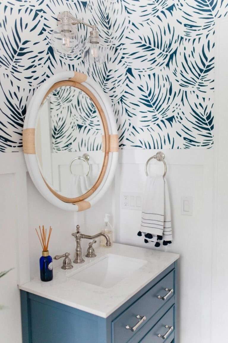 Tropical bathroom with blue palm leaf wallpaper white vanity cabinet polished gold sconce light.