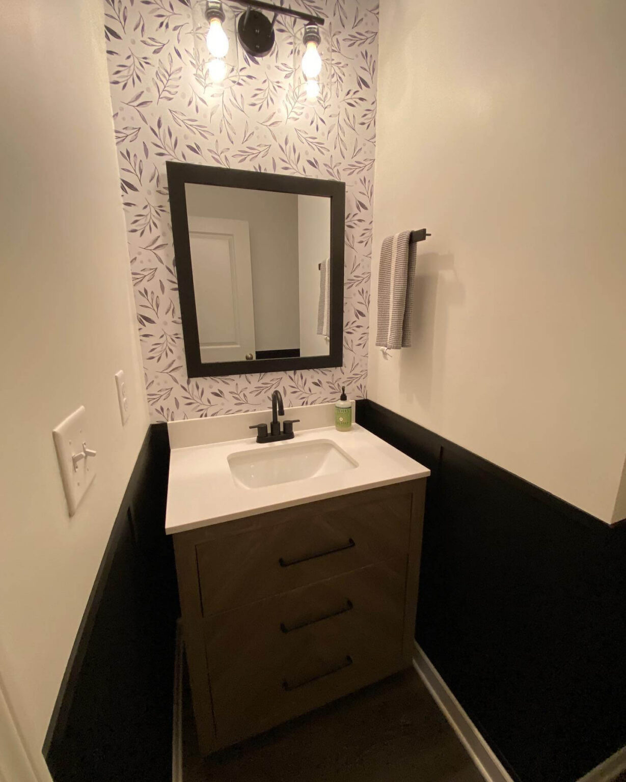 Half bathroom with black wainscoting, botanical wallpaper, white vanity, chrome fixtures.