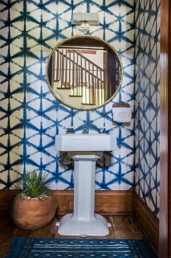 Cobalt blue geometric wallpaper framing a traditional porcelain pedestal sink with gold wall sconces