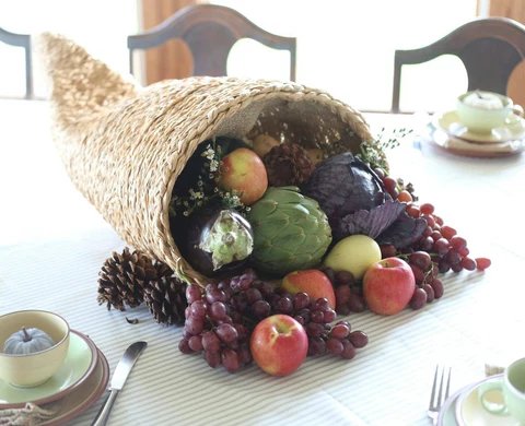 Living room coffee table, burlap cornucopia, grapes, apples, pinecones, soft amber lighting.
