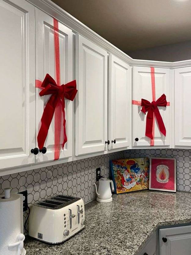 Kitchen cabinet doors covered in festive wrapping paper, topped with ribbon bows and small pinecone ornaments.