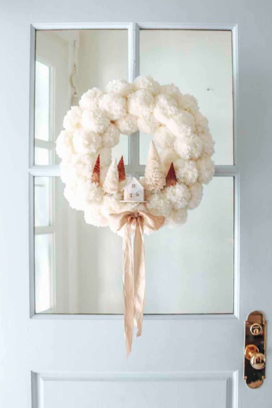 A wreath made entirely of fluffy white pom poms hanging on a neutral-colored door.