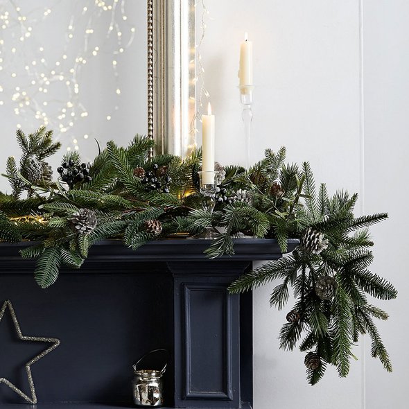 Elegant dining room table displaying lush evergreen garland, glass pillar candles, ceramics, and warm candlelight.