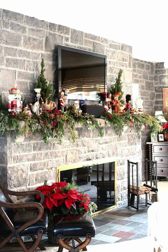 Traditional living room, TV above mantel, nutcracker figurines, evergreen garland, deep red cushions, warm lighting.