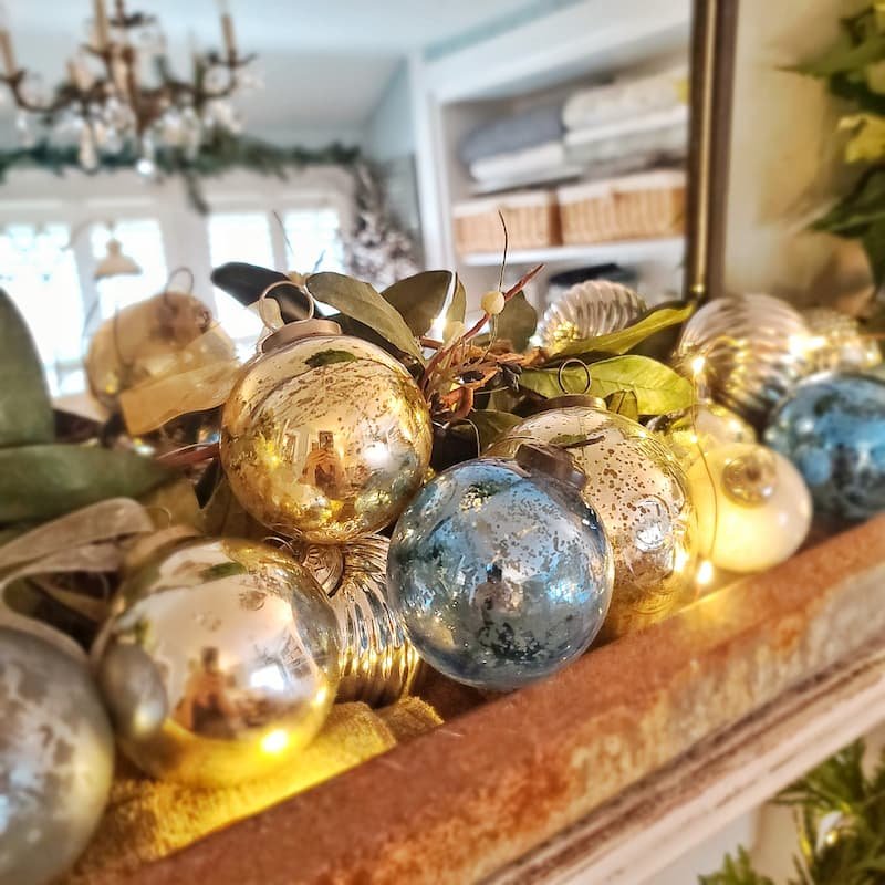 Kitchen island featuring a three-tier carved wooden tray filled with pinecones, frosted baubles, and warm lights.