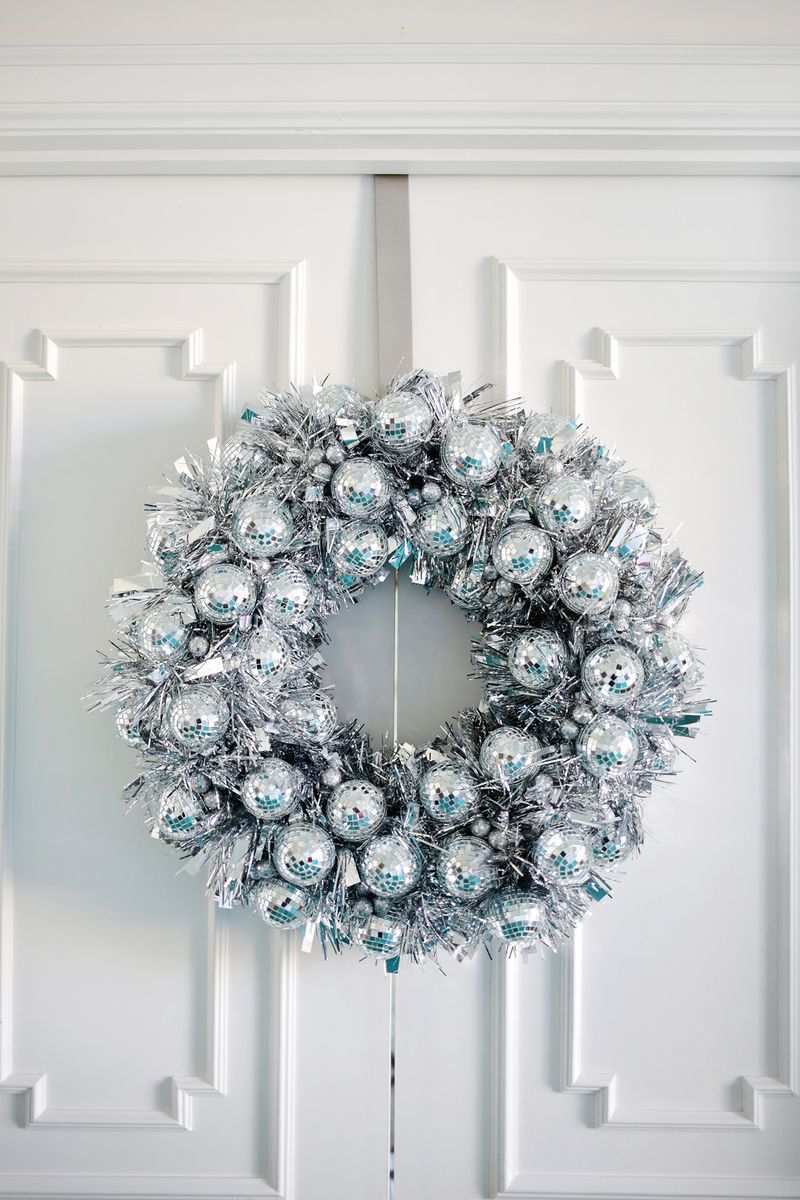 Frosted eucalyptus leaves surrounding silver and sage green christmas ornament wreath on a grey front door with ambient warm light.