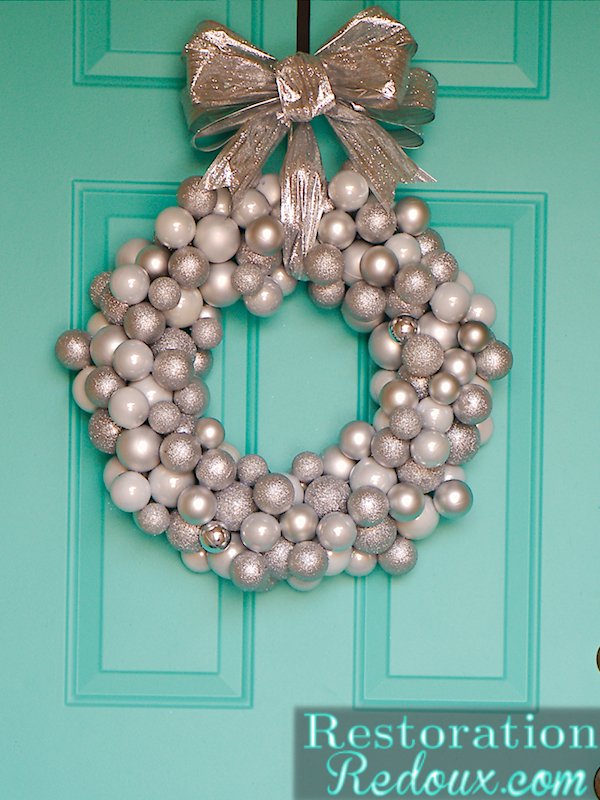 Black front door decorated with sapphire blue and silver ornament wreath, warm lantern light, and doormat.