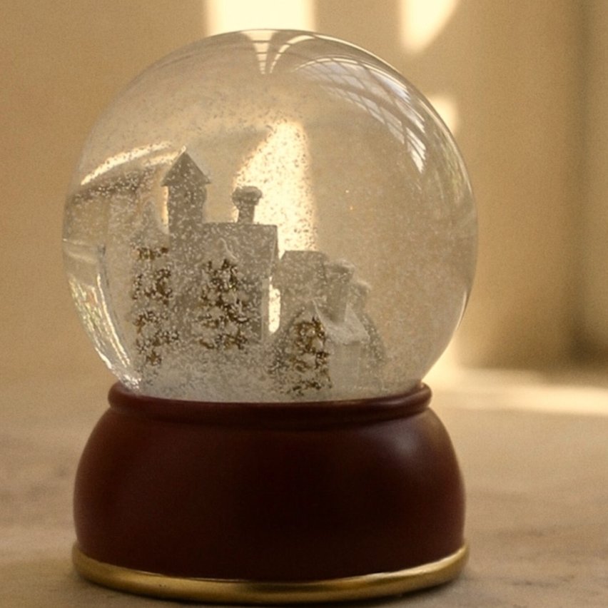 Large christmas village snow globe with lit houses and falling snow on a wooden side table.