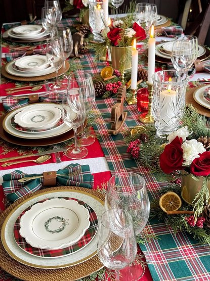 Dining room with red table runner, green foliage centerpiece, brass flatware, warm lighting.