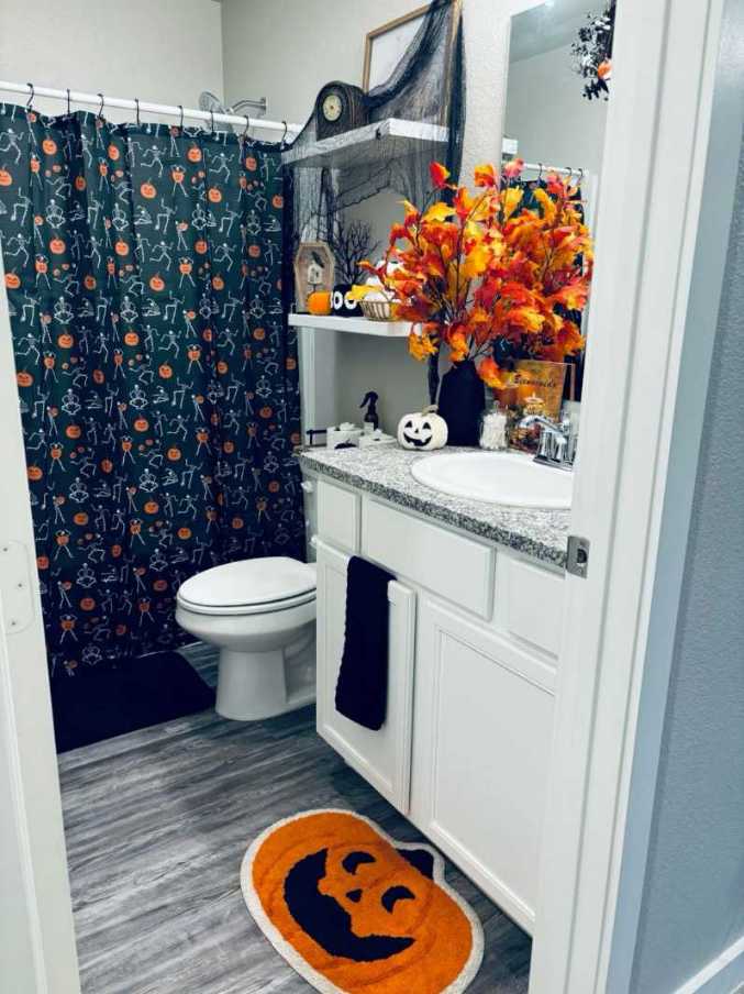 White bathroom vanity, dancing skeleton shower curtain, pumpkin rug, cool gray tiles, soft daylight.