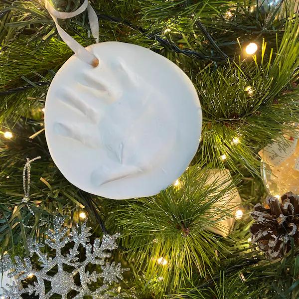 Modern holiday living room tree with a clear glass ornament bearing a tiny white baby handprint surrounded by soft neutral ribbon.