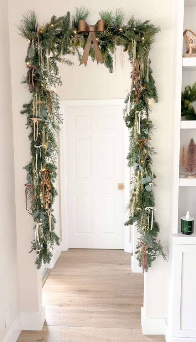 Front entry doorframe wrapped in a lush evergreen garland, interspersed with white ribbons and tiny LED lights.