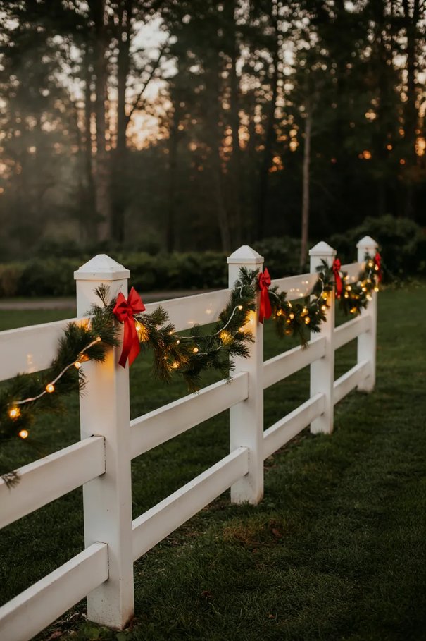 White wooden picket fence styled with fresh cedar garland, warm fairy lights, red berry accents.
