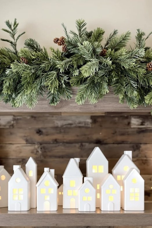 Mantel with small glowing ceramic houses, pine branches, and warm white string lights.