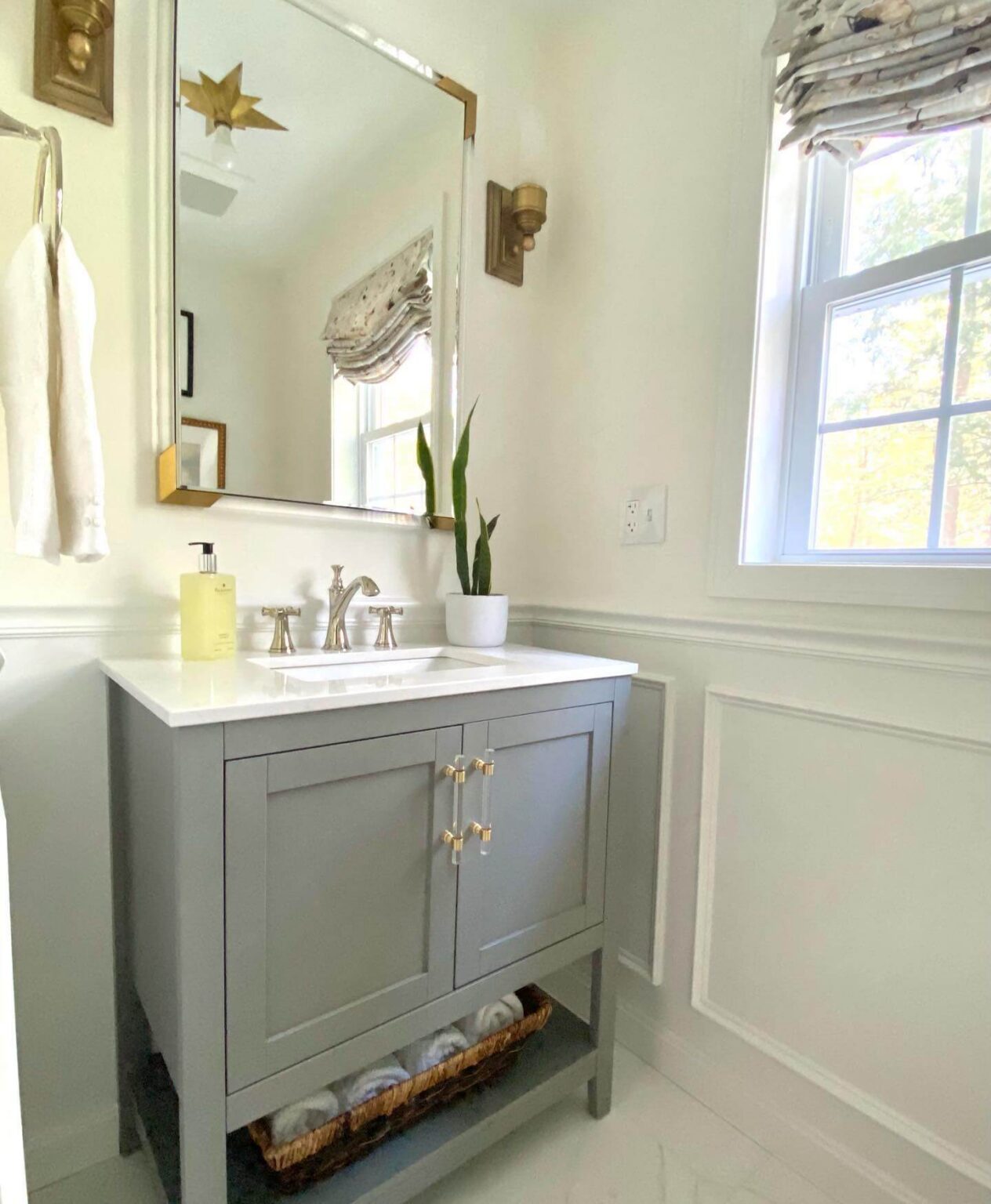 Half bathroom with grey vanity, white wainscoting, brass framed mirror, sleek fixtures.
