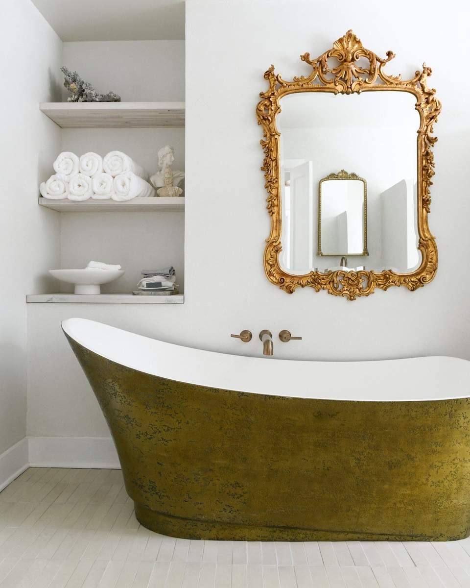 Guest bathroom with striking metallic gold clawfoot tub, ornate antique gold mirror, and warm chandelier lighting.
