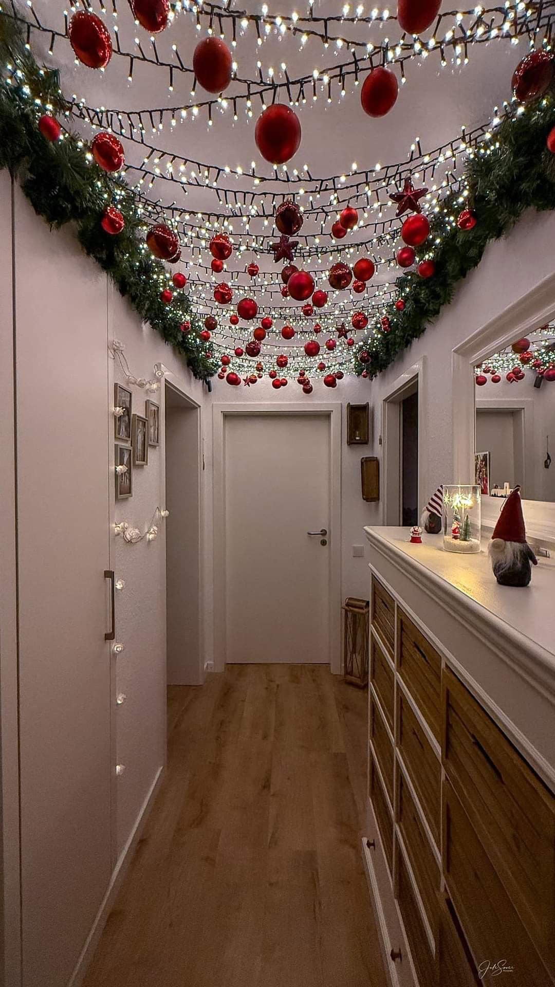 Narrow hallway ceiling adorned with a string of warm white lights and dangling glossy red baubles against light gray walls.