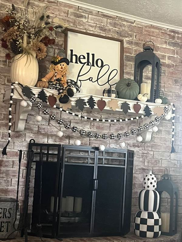 Fireplace wall, brick mantel, wooden “Hello Fall” sign, mixed autumn foliage garland, amber floor lamp.