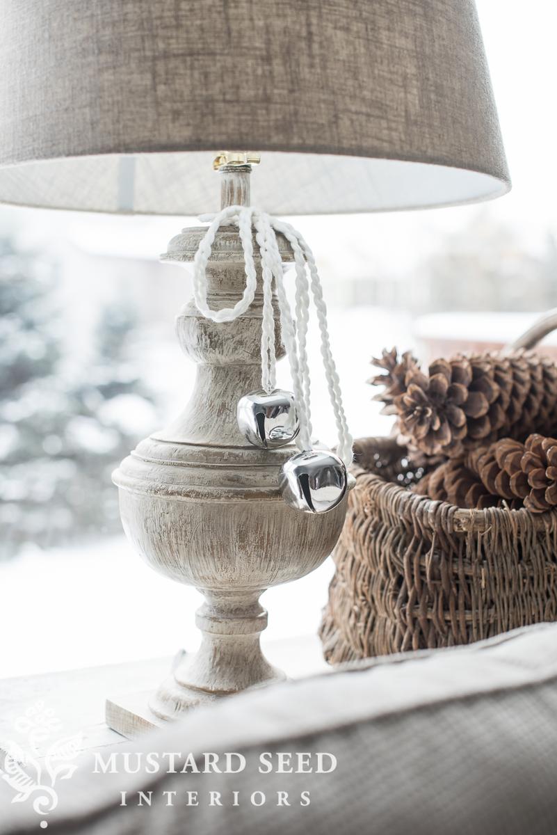 Cozy bedroom with linen curtains, cascading gold jingle bells, wood bench, and warm natural winter light.
