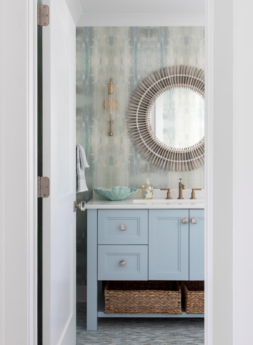 Light blue bathroom vanity with marble top and a sleek brushed brass round mirror overhead.