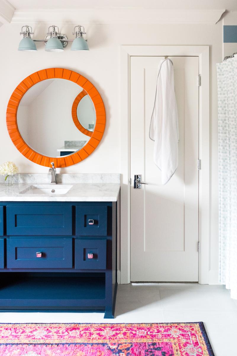 Navy vanity contrasted by a bold orange mirror and eclectic wallpaper in a chic bathroom.