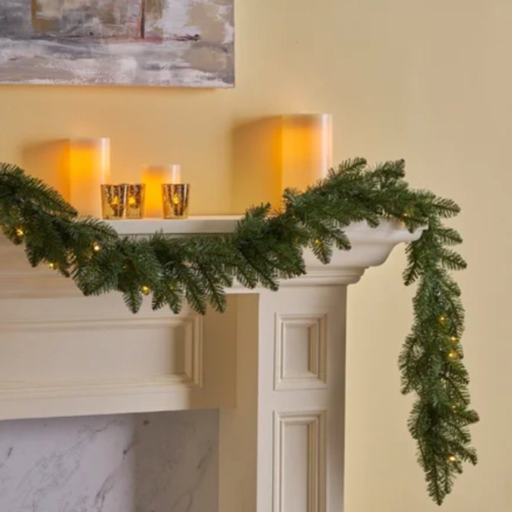 Green pre-lit mantel garland with warm lights draped across a white mantel with red bows.