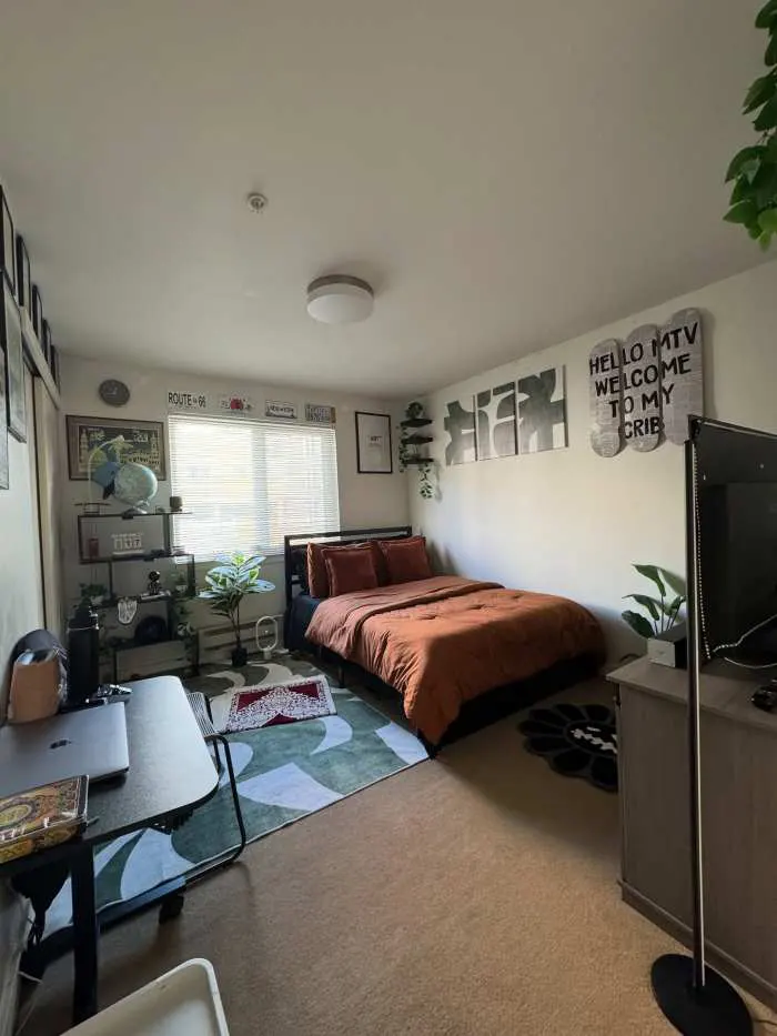 Rust-colored bedding framed by large apartment windows, offering an industrial yet cozy bedroom setting.