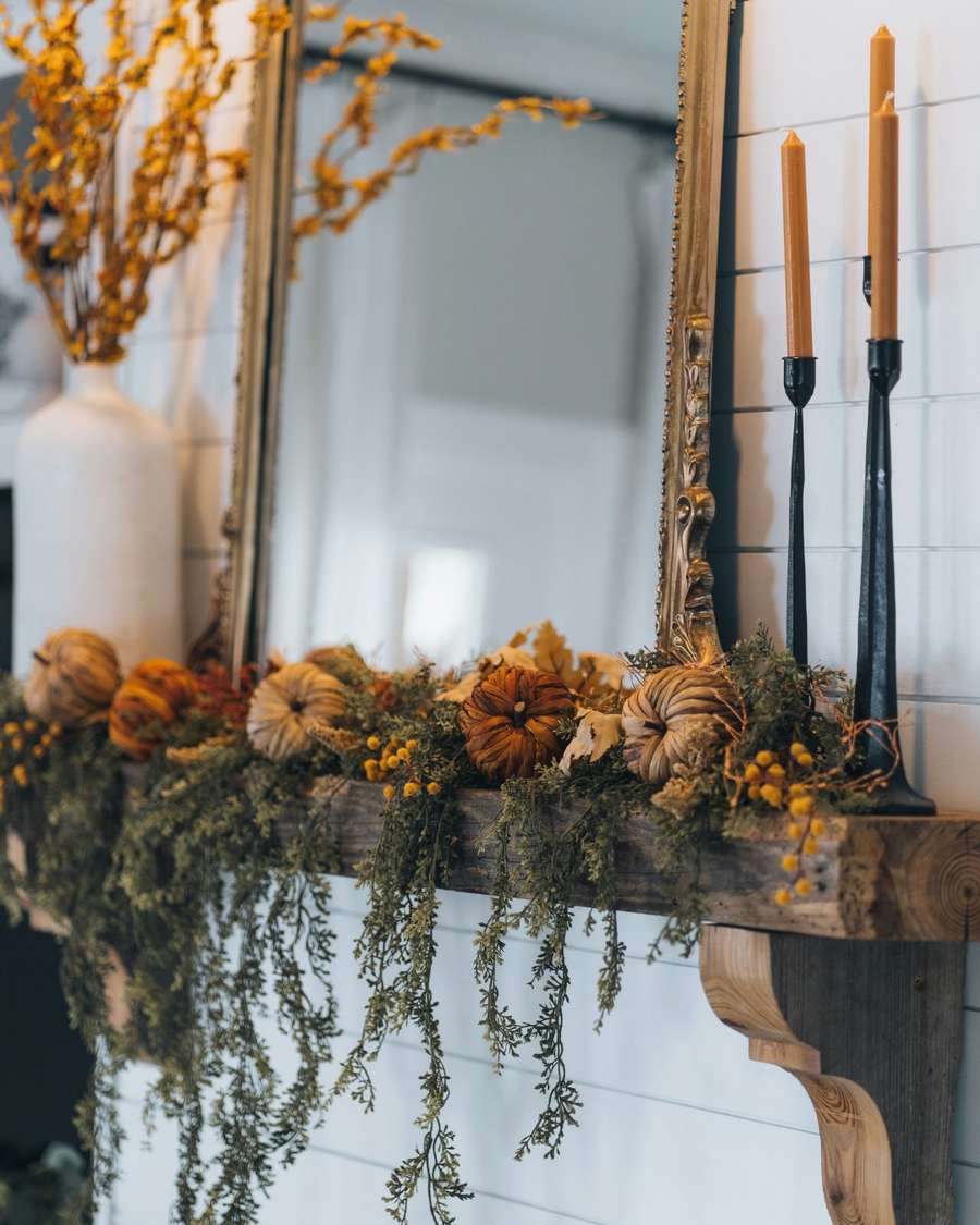 Living room wall, reclaimed wood mantel shelf, pumpkin garland, brass sconces, natural daylight.