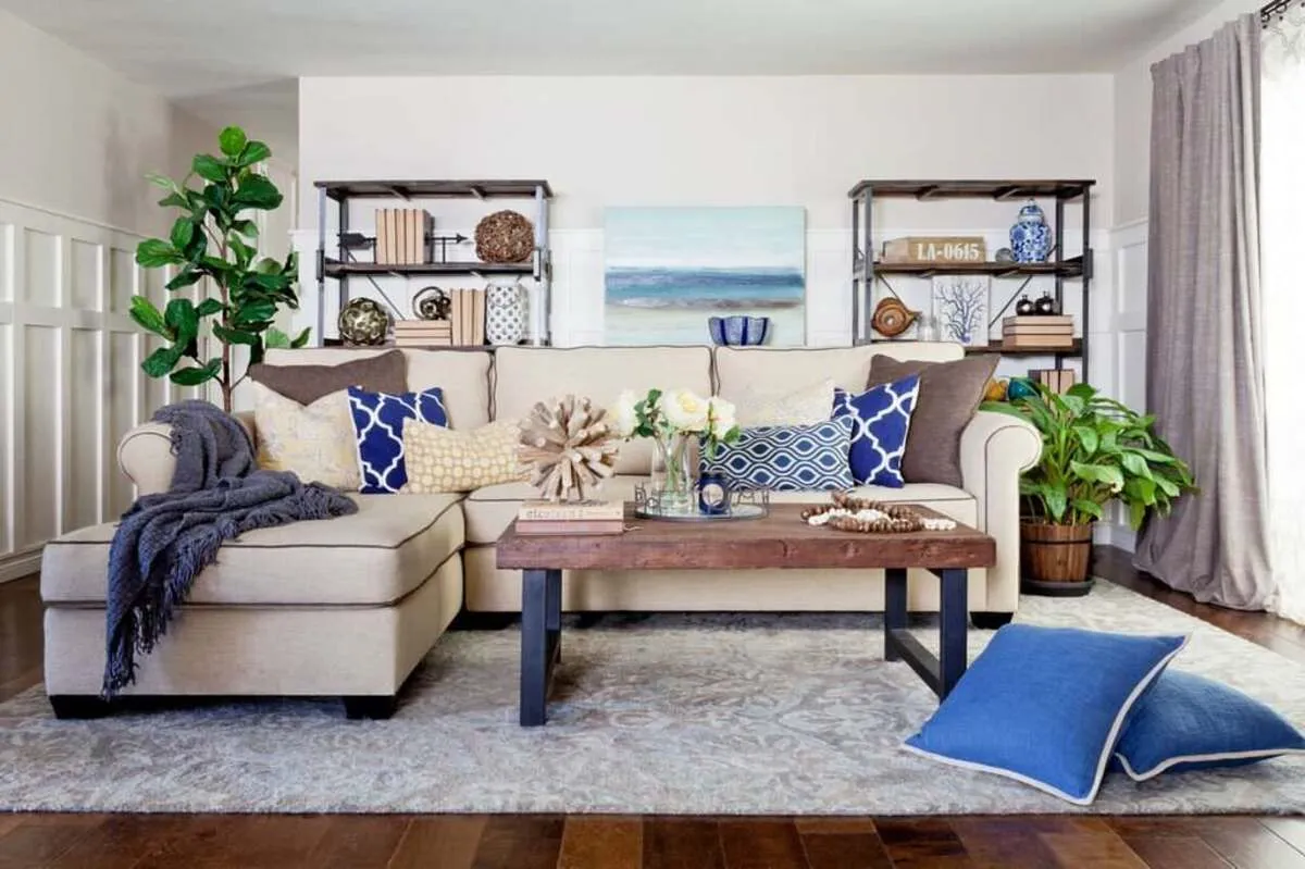 Small living room with beige sectional, blue throw pillows, green plants, and soft natural light from a large window.