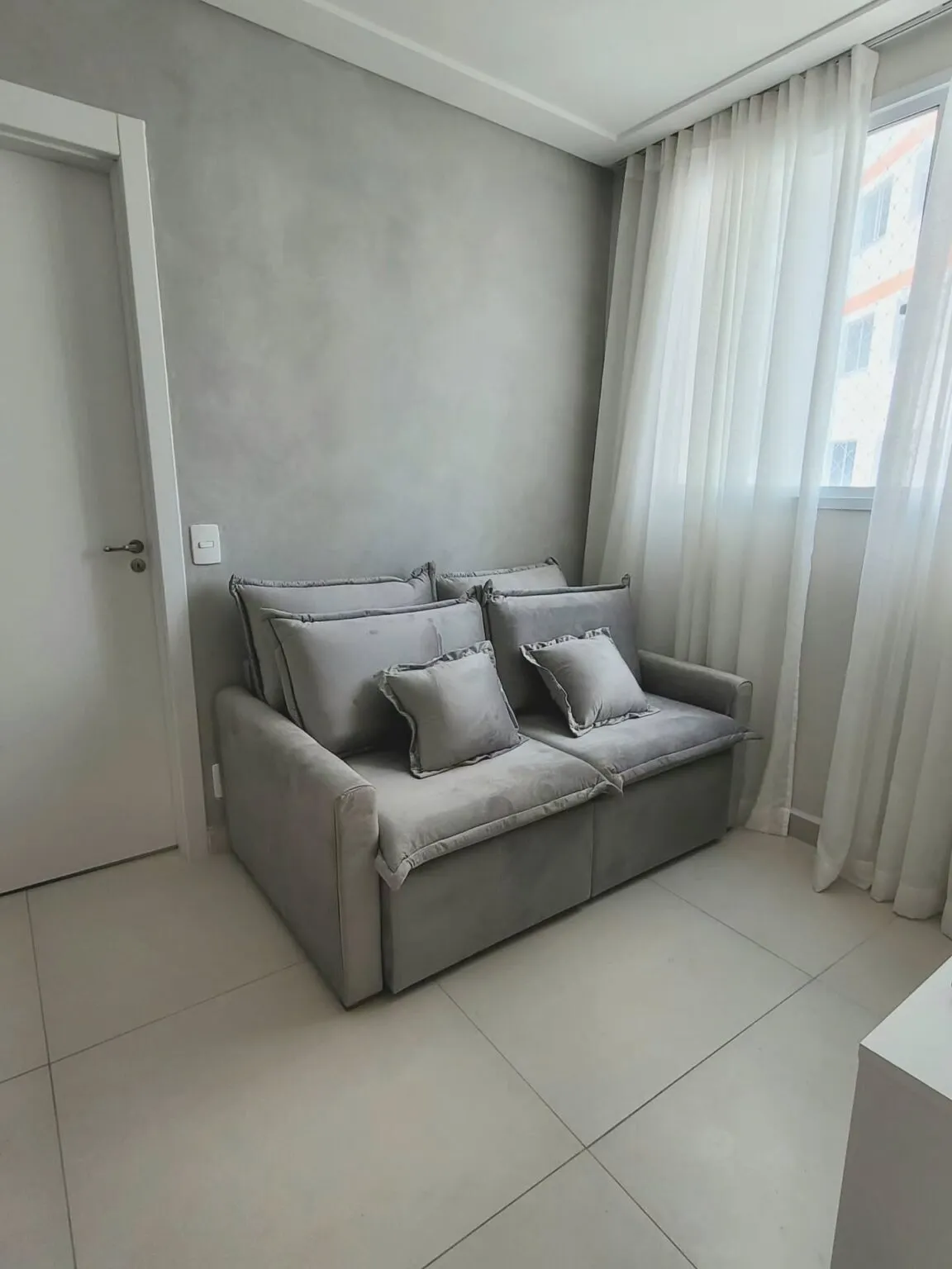 Small studio apartment living area with compact grey loveseat, round rug, and minimalist decor in soft natural light.