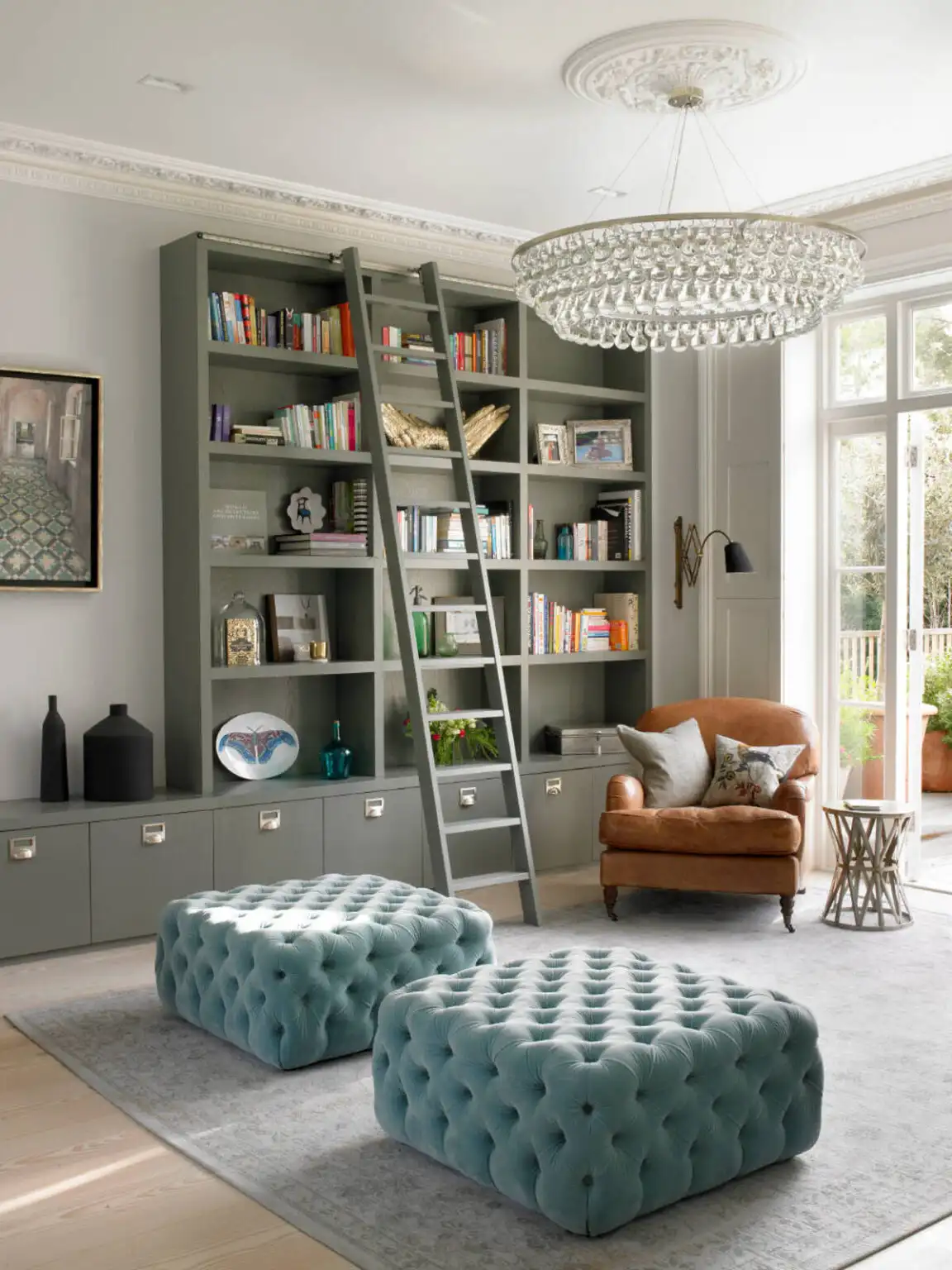 Small living room with tall bookshelves, library ladder, and tufted ottomans in warm ambient lighting.