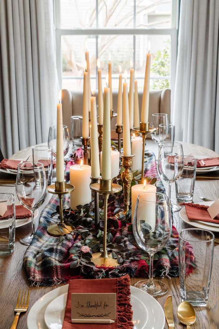 Dining table with plaid runner, three tall ivory taper candles in brass holders, warm lighting