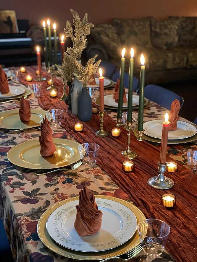 Dining table with rust-colored folded napkins, three taper candles, patterned floral tablecloth, warm lighting