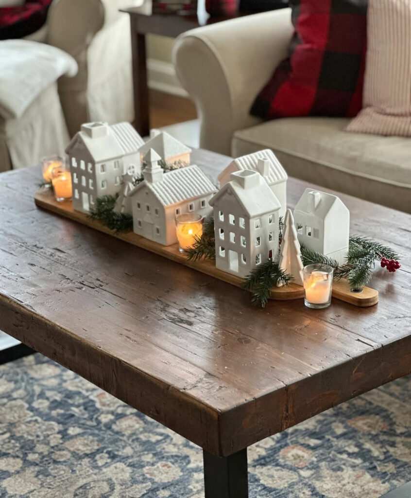 White ceramic house tealight holders arranged with pine sprigs for cozy Christmas coffee table decor.