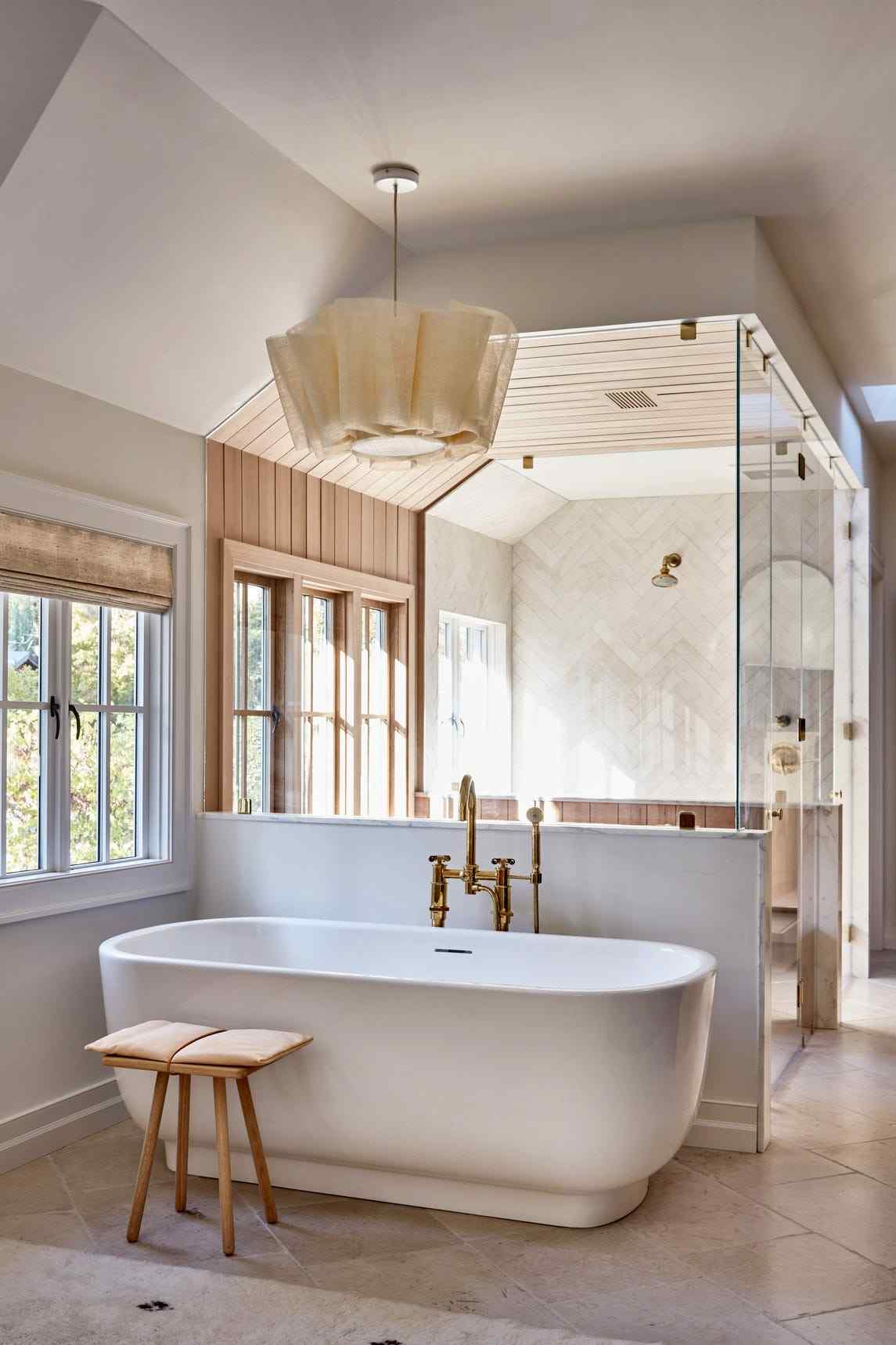 Modern white bathroom featuring oval freestanding tub, floor-mounted brass faucet, marble floors, and sunny window