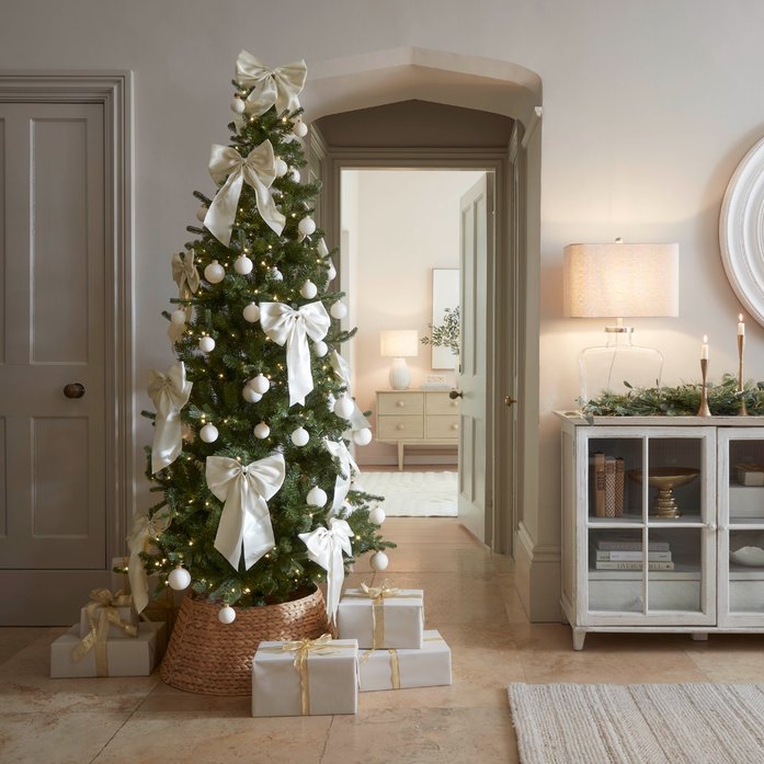 Petite apartment corner with white bow slim christmas tree beside velvet accent chair in soft lighting
